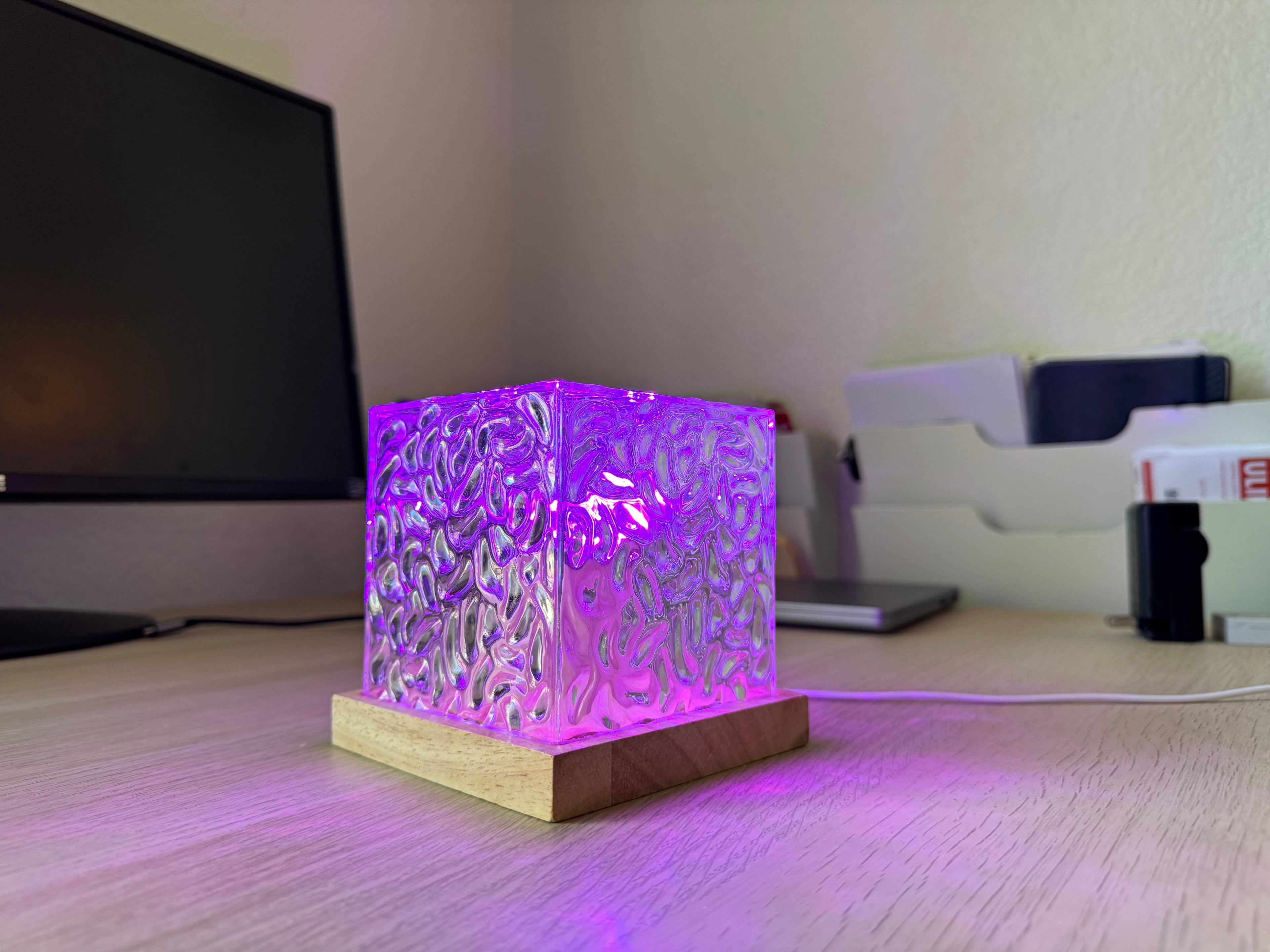 A square LED light box with a textured, frosted acrylic front and a light wood base, glowing with purple light on a desk next to a computer monitor.