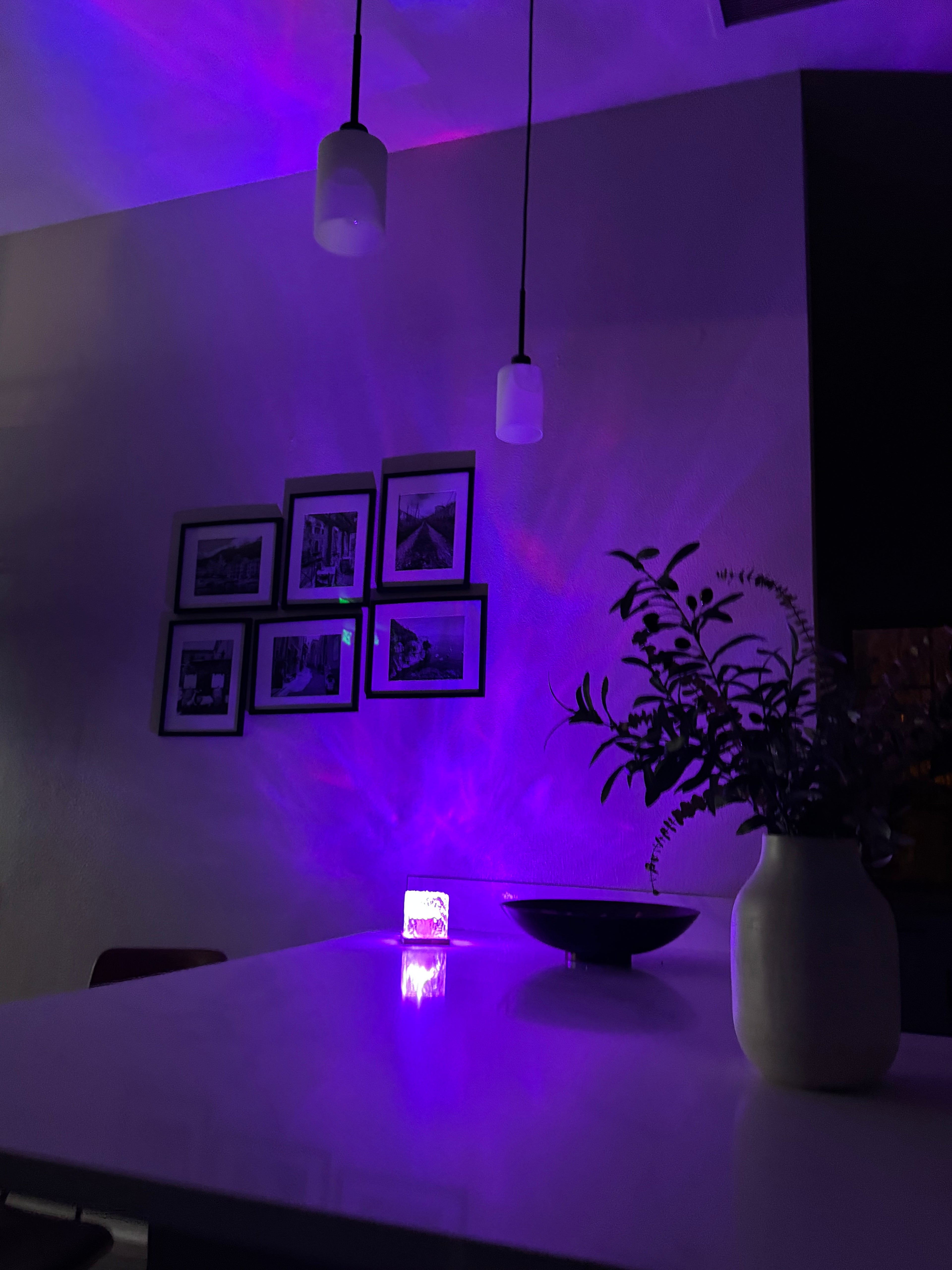 A purple LED wall light casting light on a wall with six framed black-and-white photos, a hanging plant, and pendant lights in a room setting.